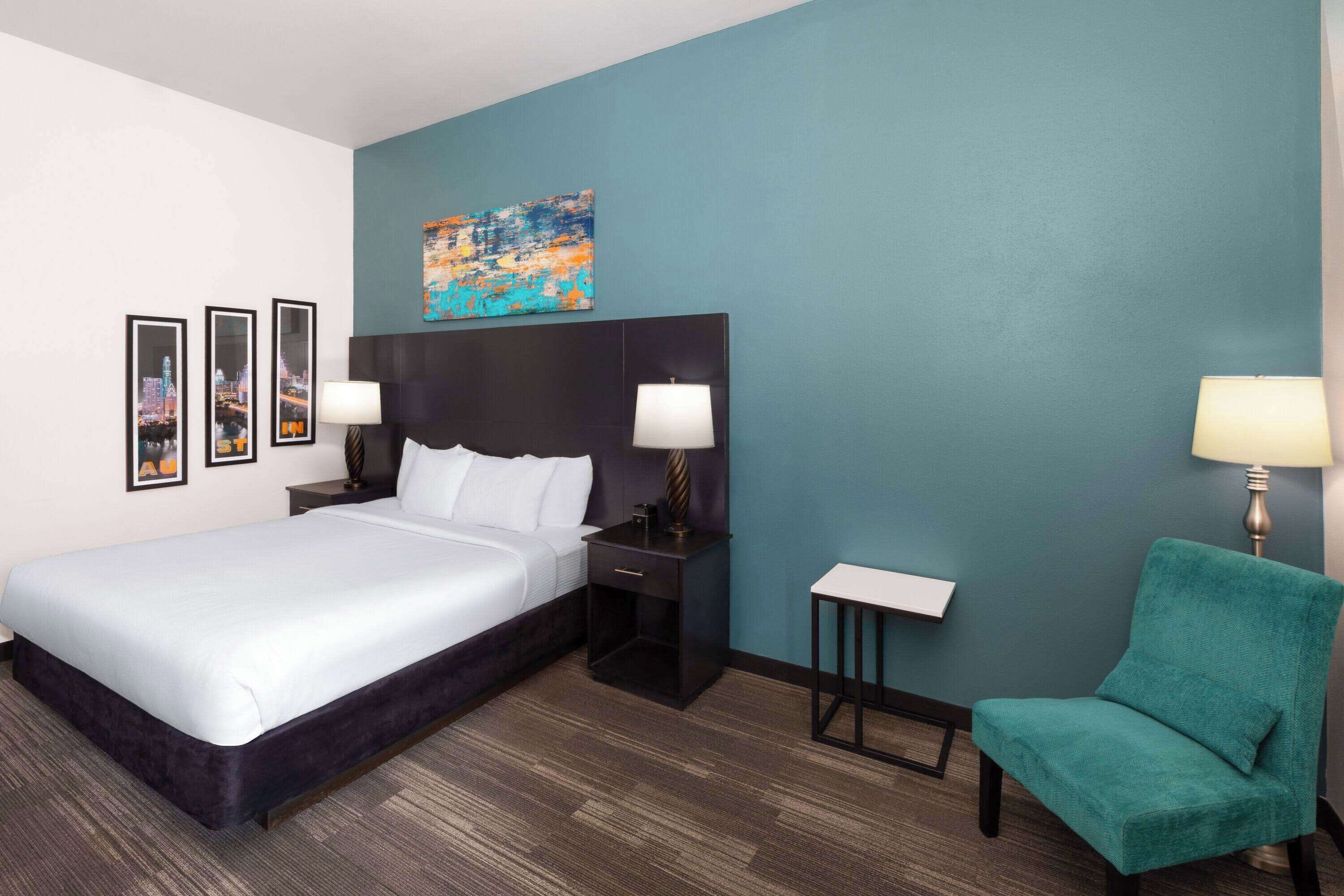 A hotel room with a large bed, a headboard, two bedside tables with lamps, a small side table, a teal armchair, and a cityscape painting on the wall.