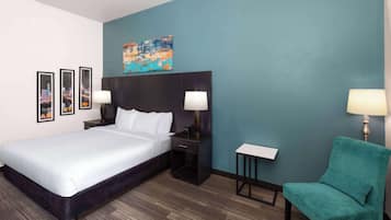 A hotel room with a large bed, a headboard, two bedside tables with lamps, a small side table, a teal armchair, and a cityscape painting on the wall.