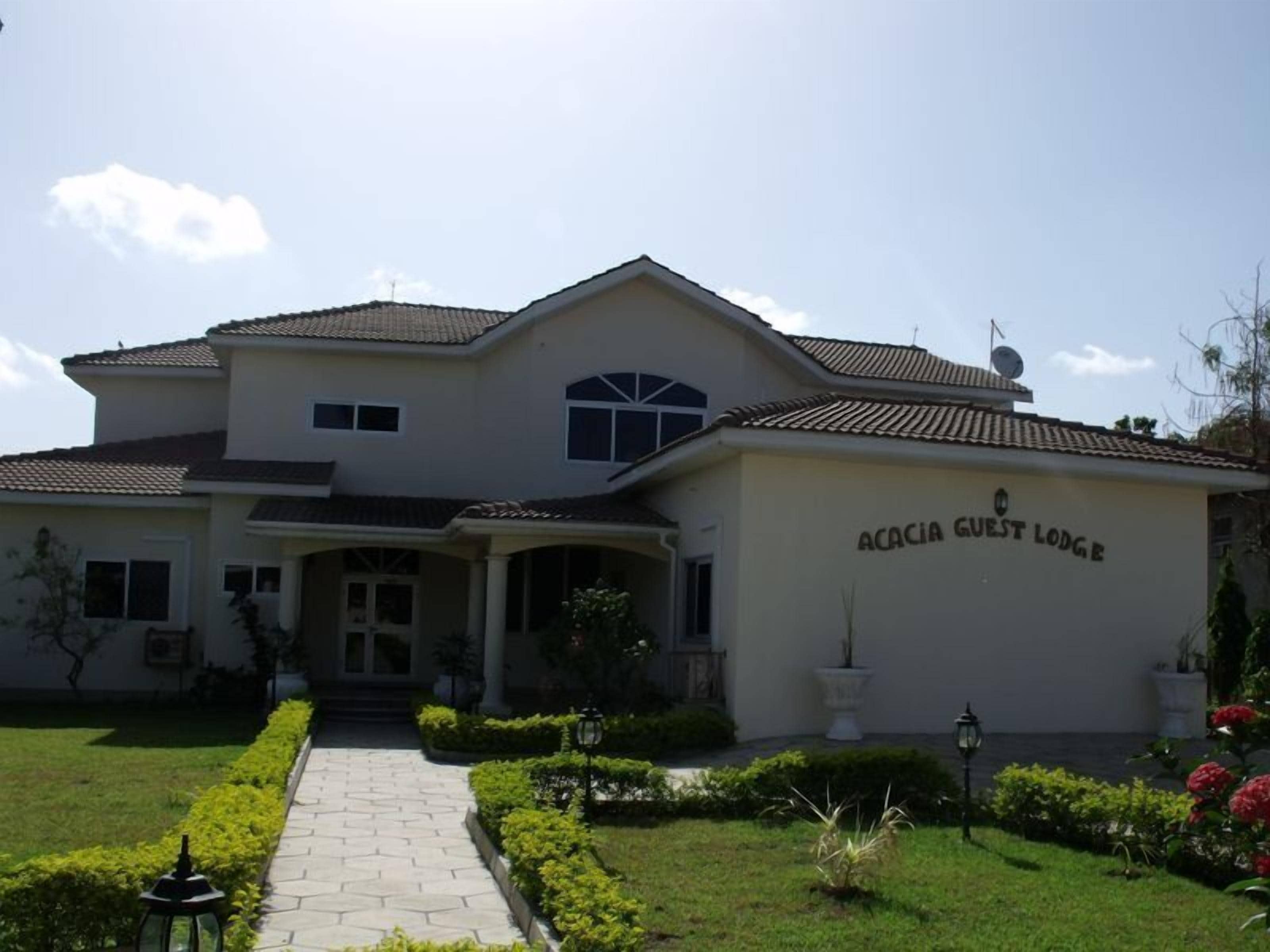 A two-story building labeled "ACACIA GUEST LODGE" with a tiled roof, a central entrance, and a landscaped garden with a pathway leading to the entrance.