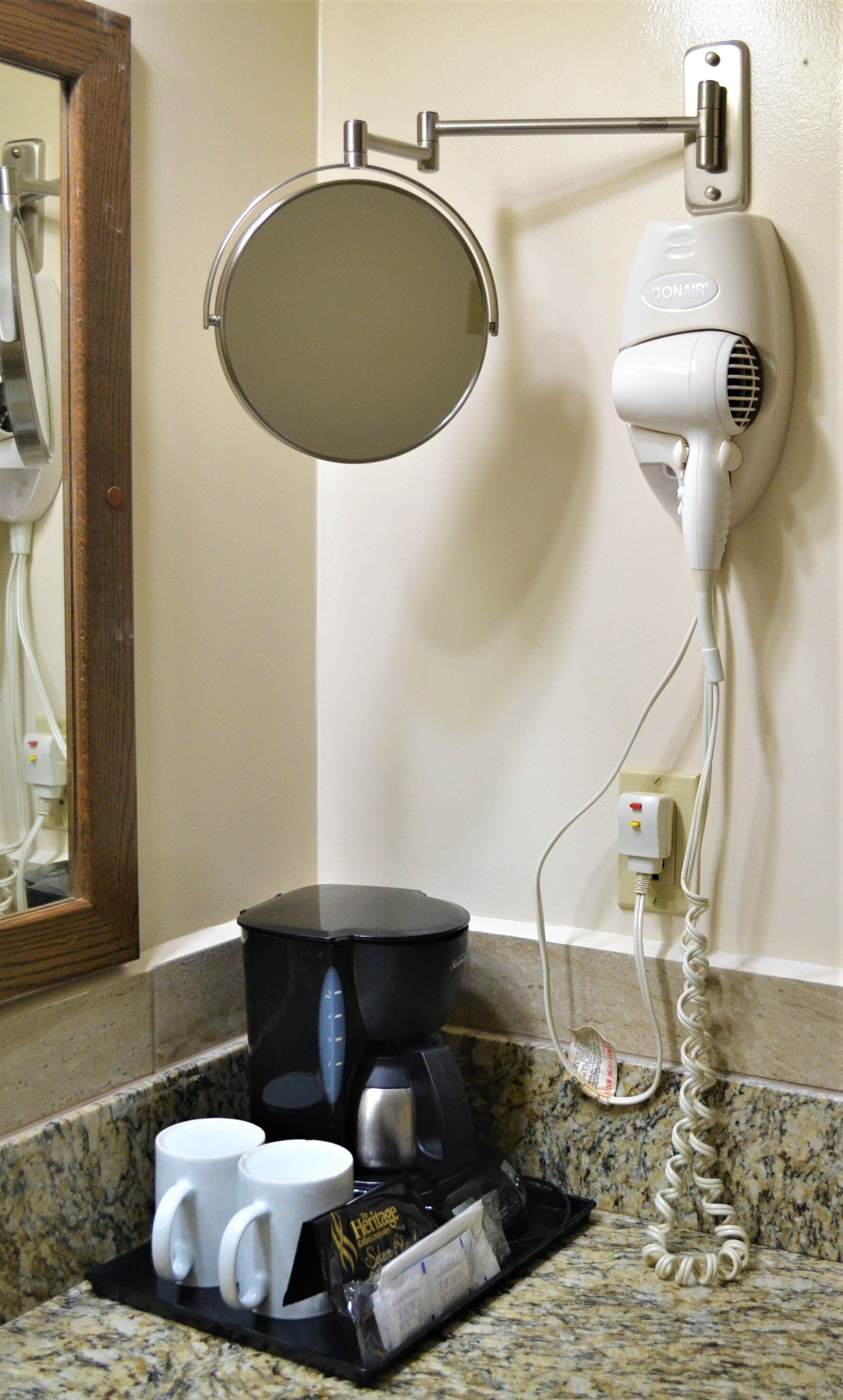 A bathroom with a hairdryer, a round mirror, a coffee maker, and two white mugs on the counter.