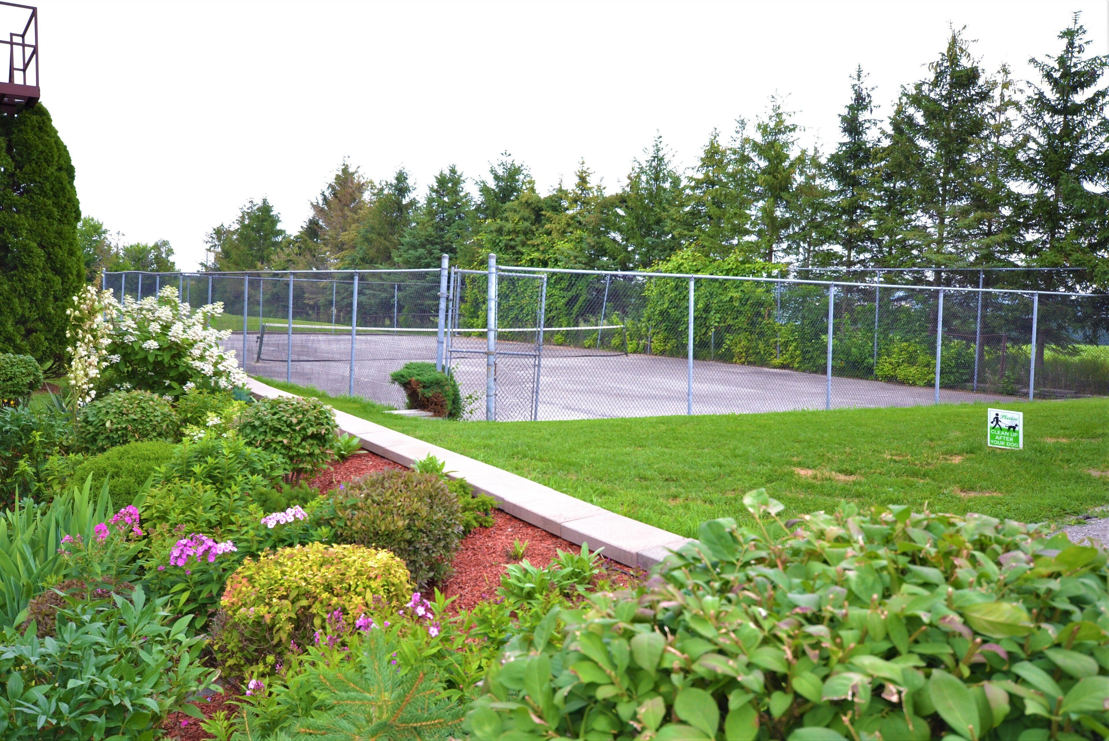 A fenced outdoor area with a paved walkway, surrounded by lush greenery and various flowering plants.