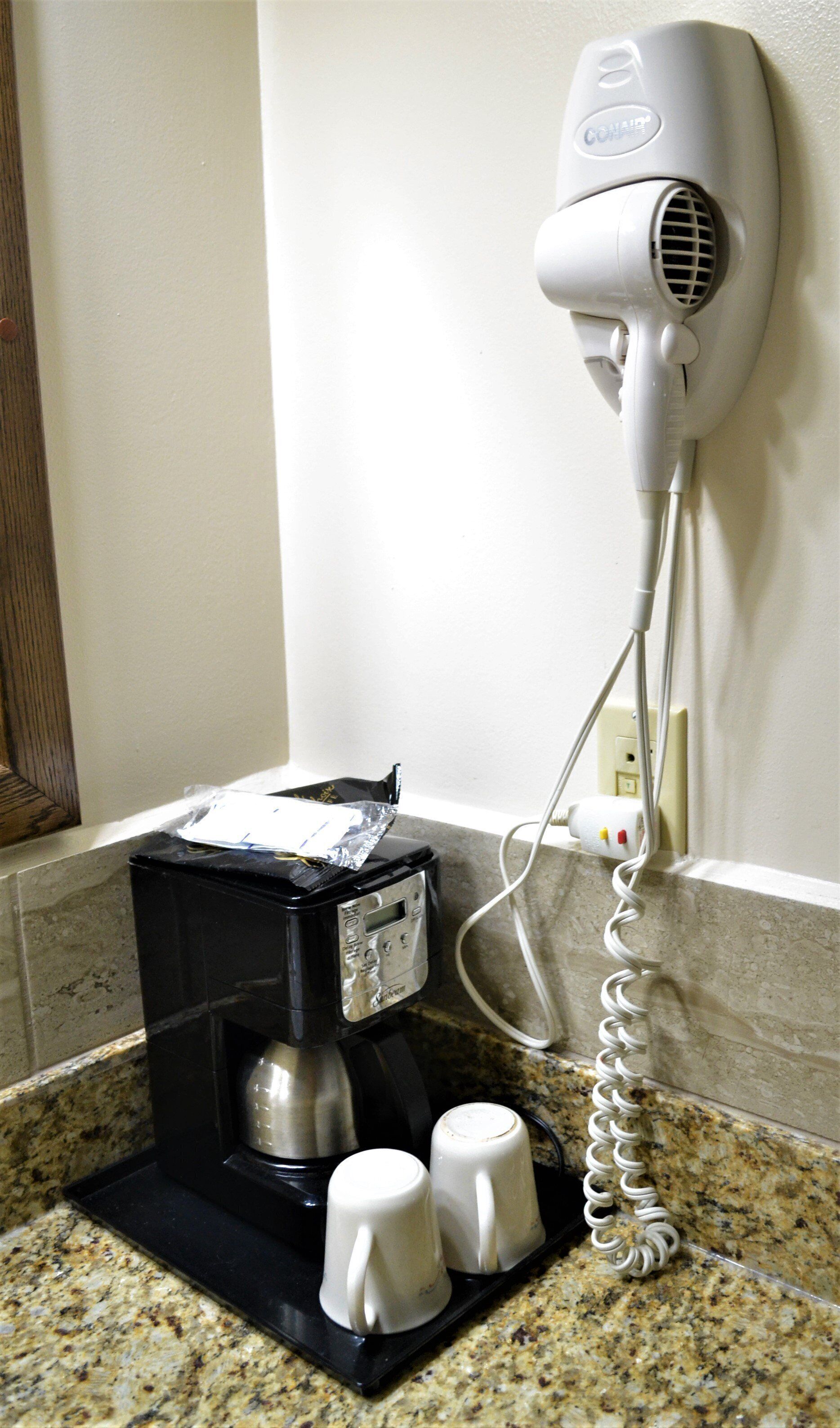 A coffee maker with two white mugs on a granite countertop, a hair dryer on the wall, and a power outlet.