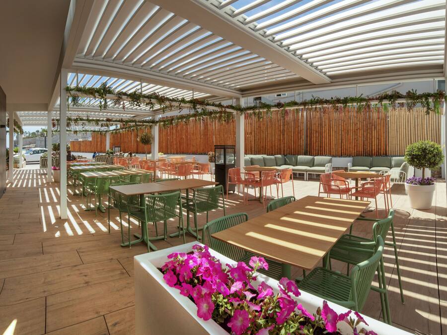 Un café en plein air avec des tables et des chaises en bois, un plancher en bois et un espace de repos couvert, délimité par une clôture en bambou.