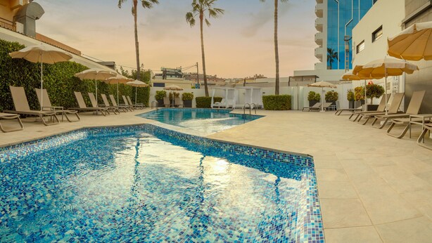 Una piscina con bordes de azulejos, rodeada de sillones de descanso y sombrillas, con palmeras y un edificio moderno al fondo.