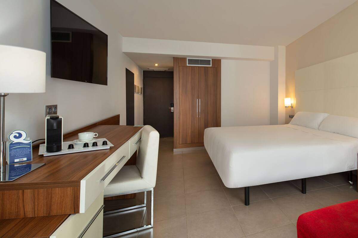 Habitació d'hotel amb llit, escriptori amb ordinador, cadira, sofà vermell i televisió penjada a la paret.