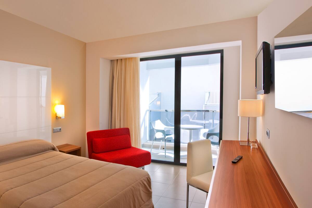 Habitació d'hotel amb un llit, una butaca vermella, un escriptori i vista al balcó.