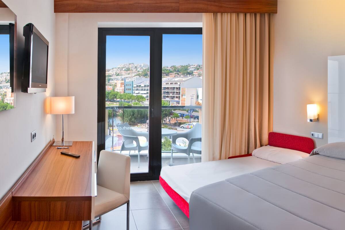 Habitació d'hotel amb llit, escriptori, cadira, televisió i un balcó amb vista a la ciutat.