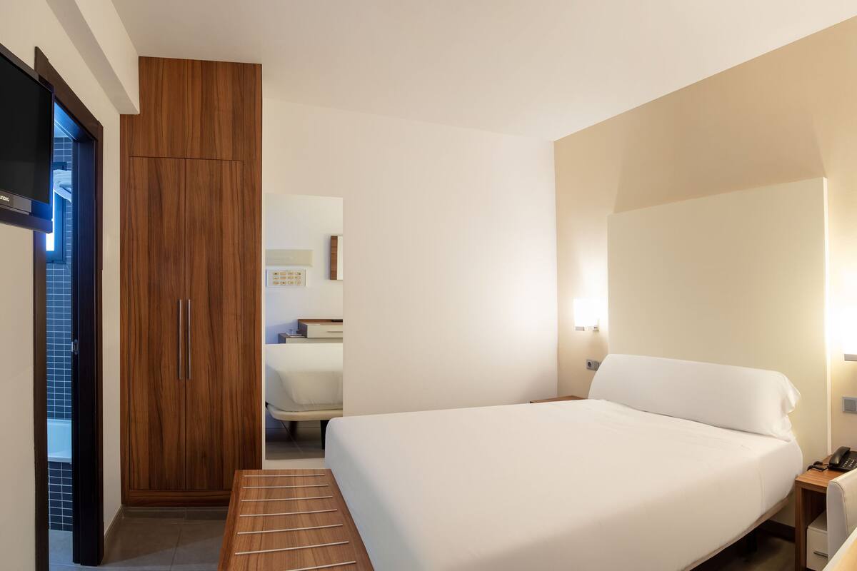 Habitació d'hotel amb llit, taula de nit, làmpada i armari de fusta.