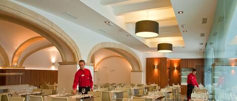 Un restaurante espacioso con techos altos, arcos y moderna iluminación colgante. Las mesas están dispuestas con copas de vino y cubiertos.