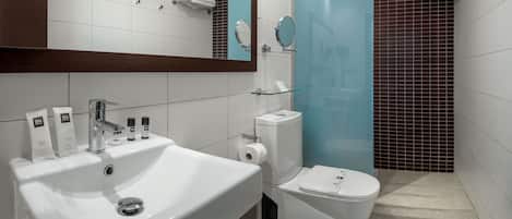 Un baño moderno con un lavabo blanco, espejo y retrete. Hay artículos de aseo en el lavabo y un estante de vidrio sobre el retrete.