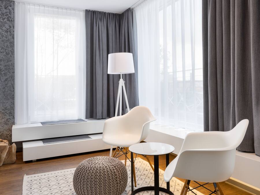 A modern living room with a white sofa, two white chairs, a small round ottoman, a floor lamp, and a window with grey curtains.