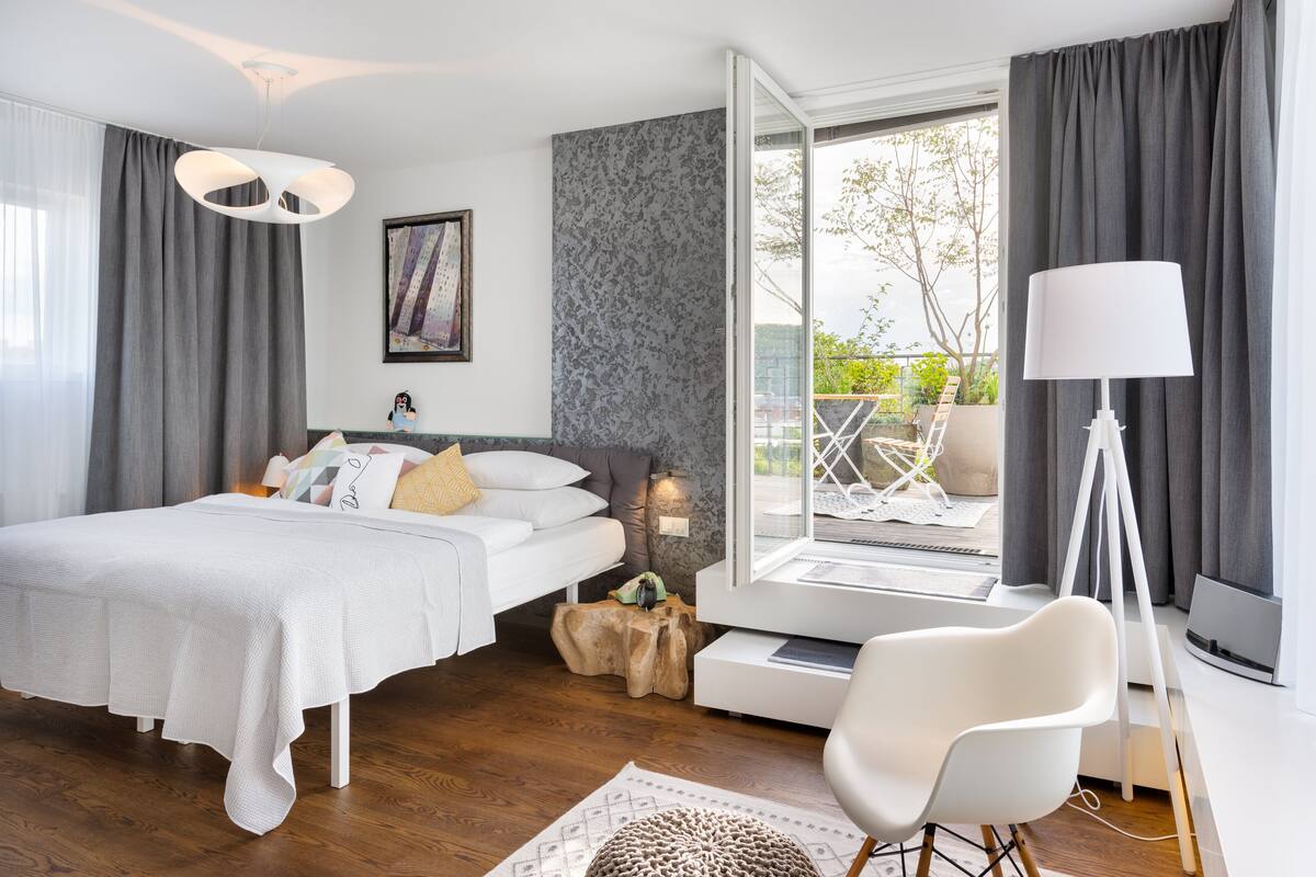 A modern bedroom with a bed, a white chair, a desk with a lamp, and a view of the outdoors.