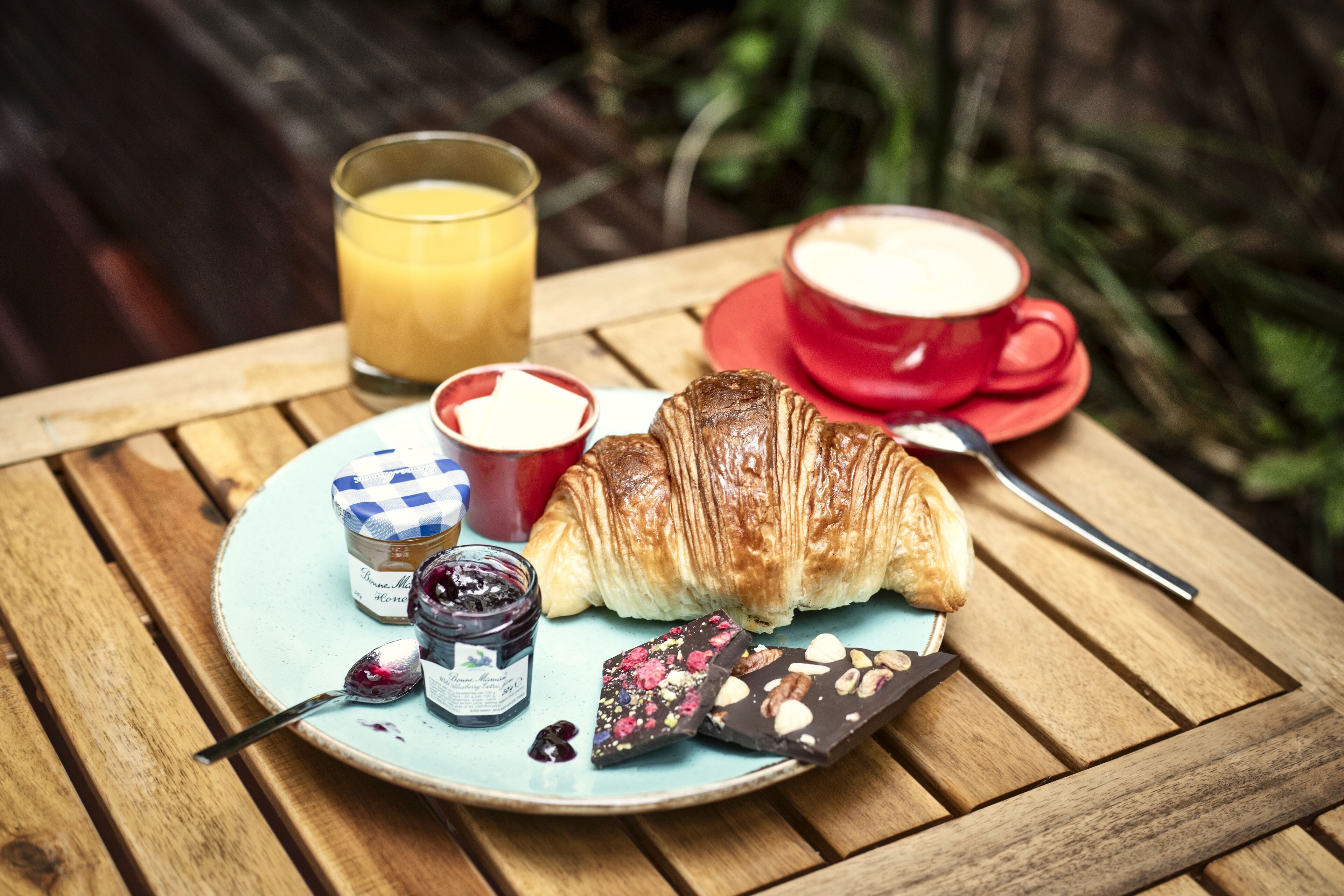 A breakfast setting with a croissant, a glass of orange juice, a cup of coffee, a small jar of jam, and a chocolate bar on a round table.