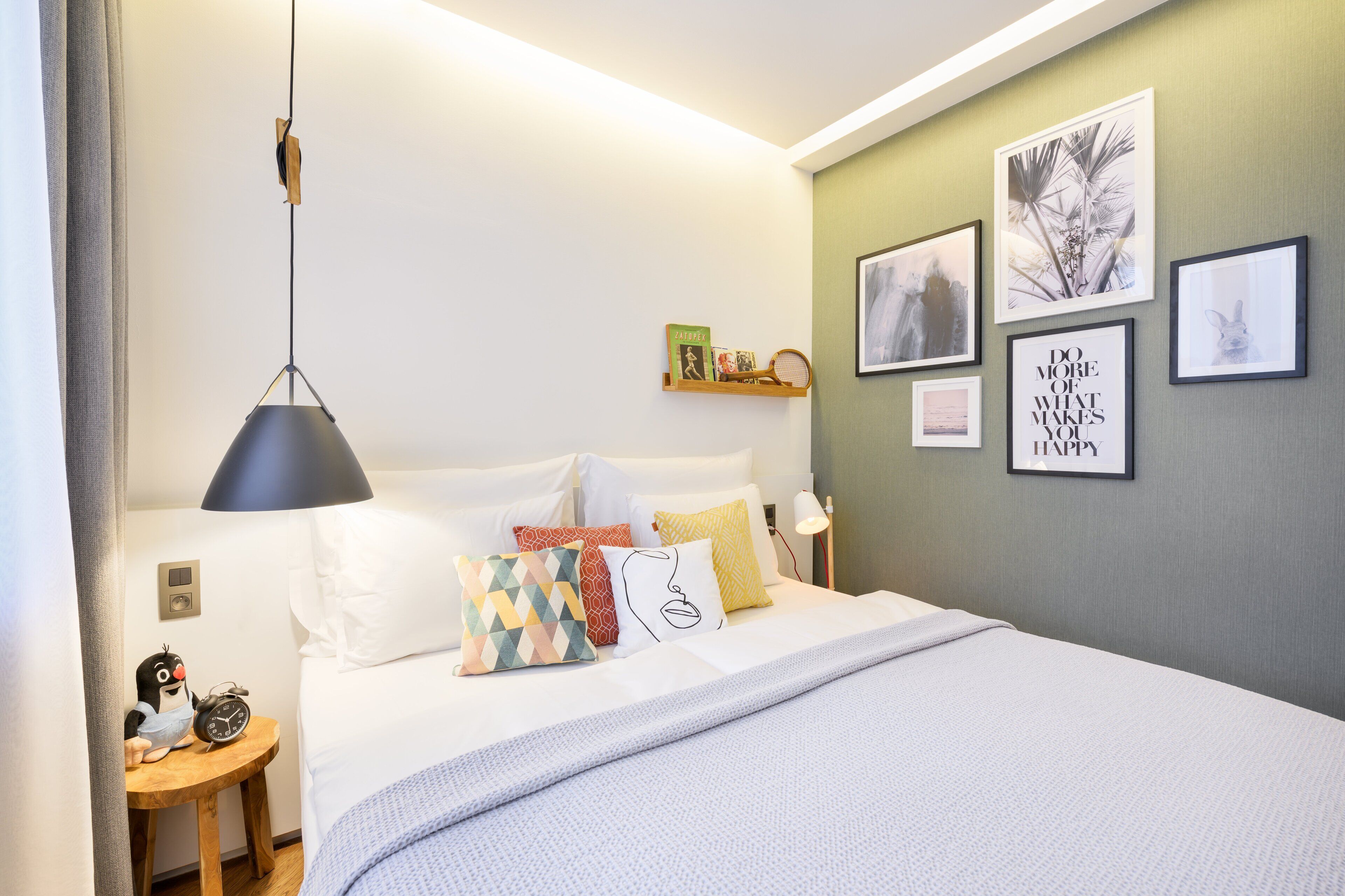 Un dormitorio ordenado con una cama, almohadas, una mesita de noche con un reloj y decoración en la pared.