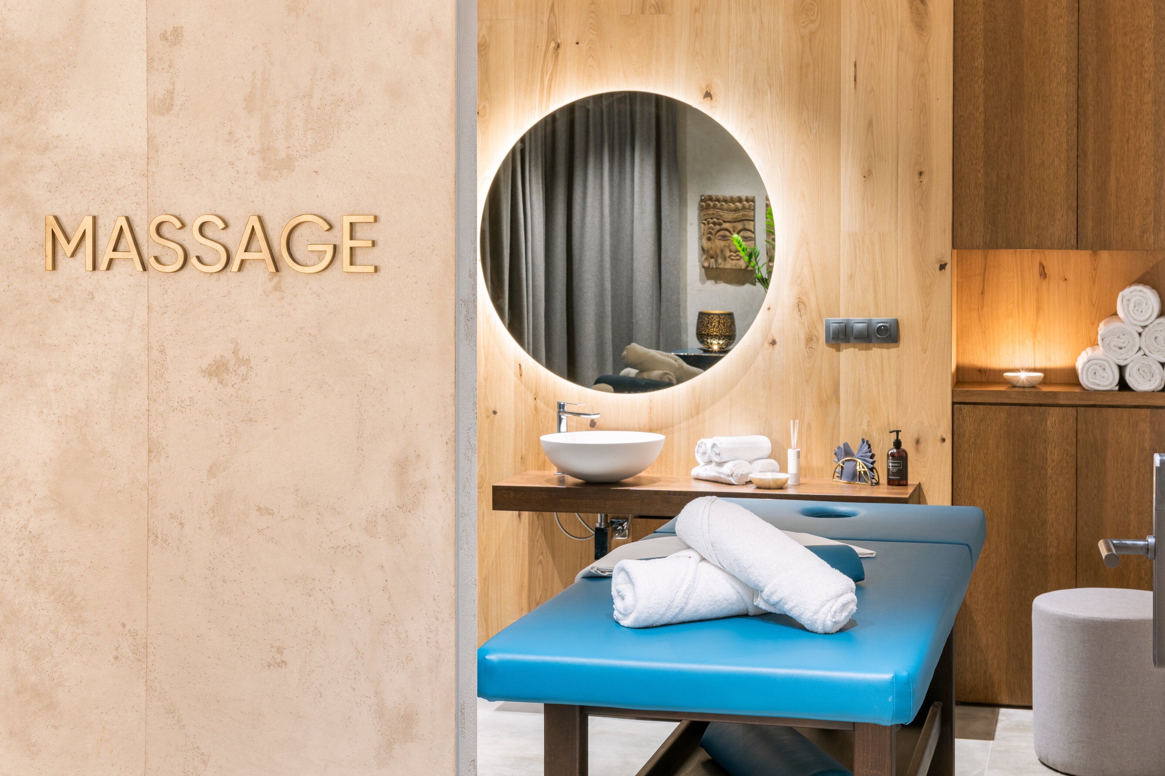 A massage room with a wooden wall, a round mirror, a blue massage table, and a sink with towels.