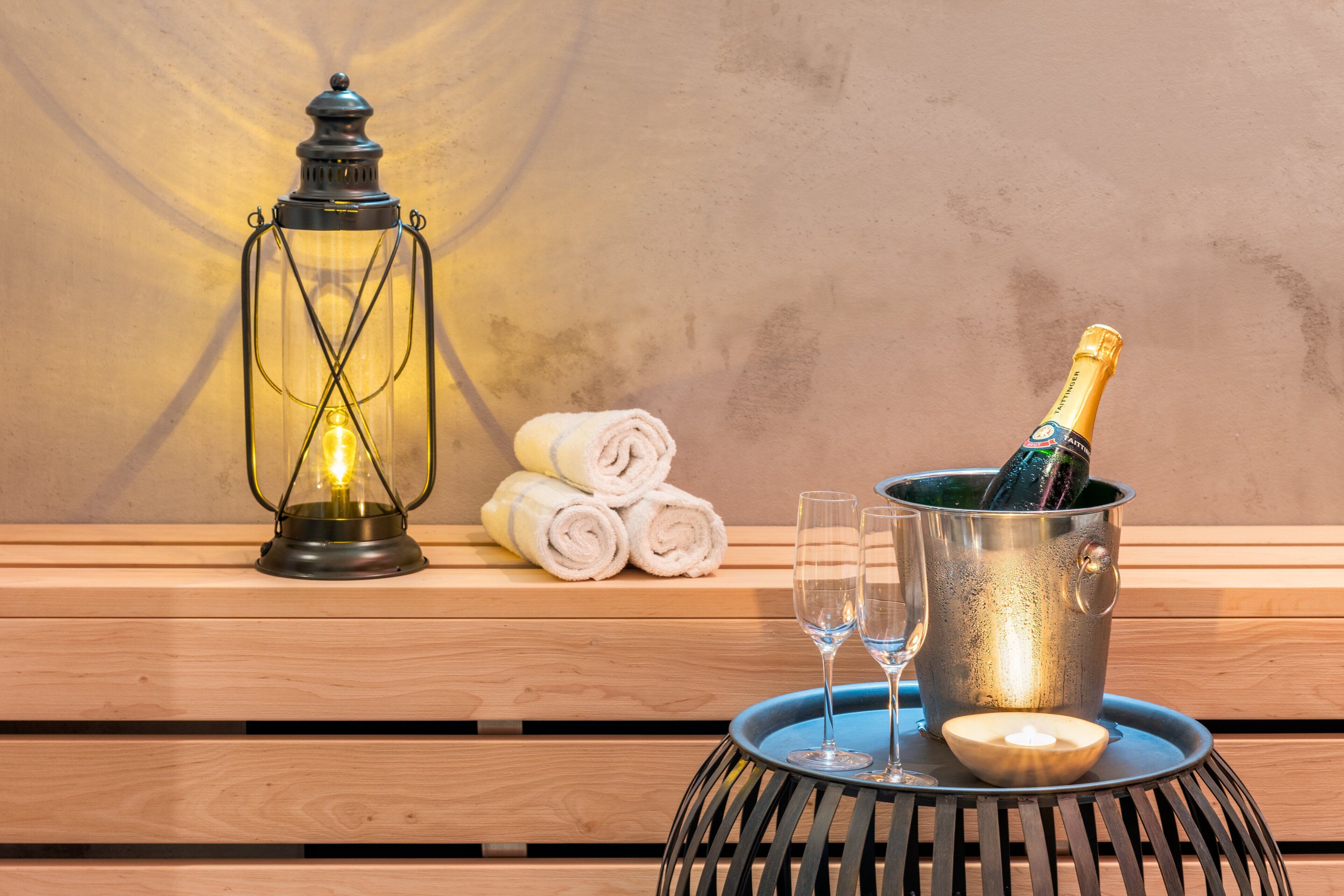 A wooden bench with a metal lantern, a bottle of champagne, two glasses, and rolled towels.
