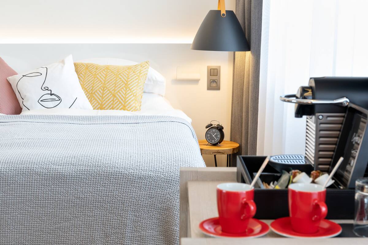 A hotel room with a bed, a bedside table with a clock, a lamp, and a tray with red cups and a glass.