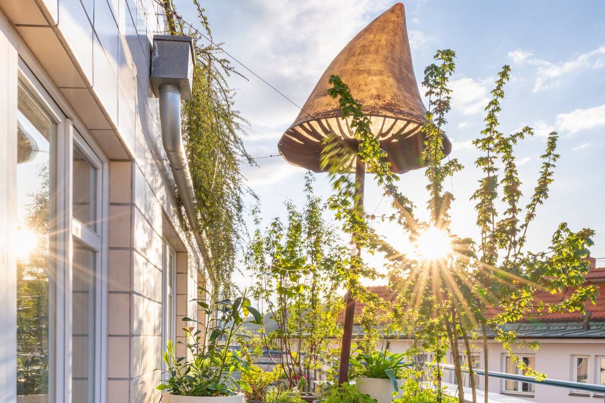 A rooftop terrace with a wooden table and benches, surrounded by potted plants and a large conical shade structure.