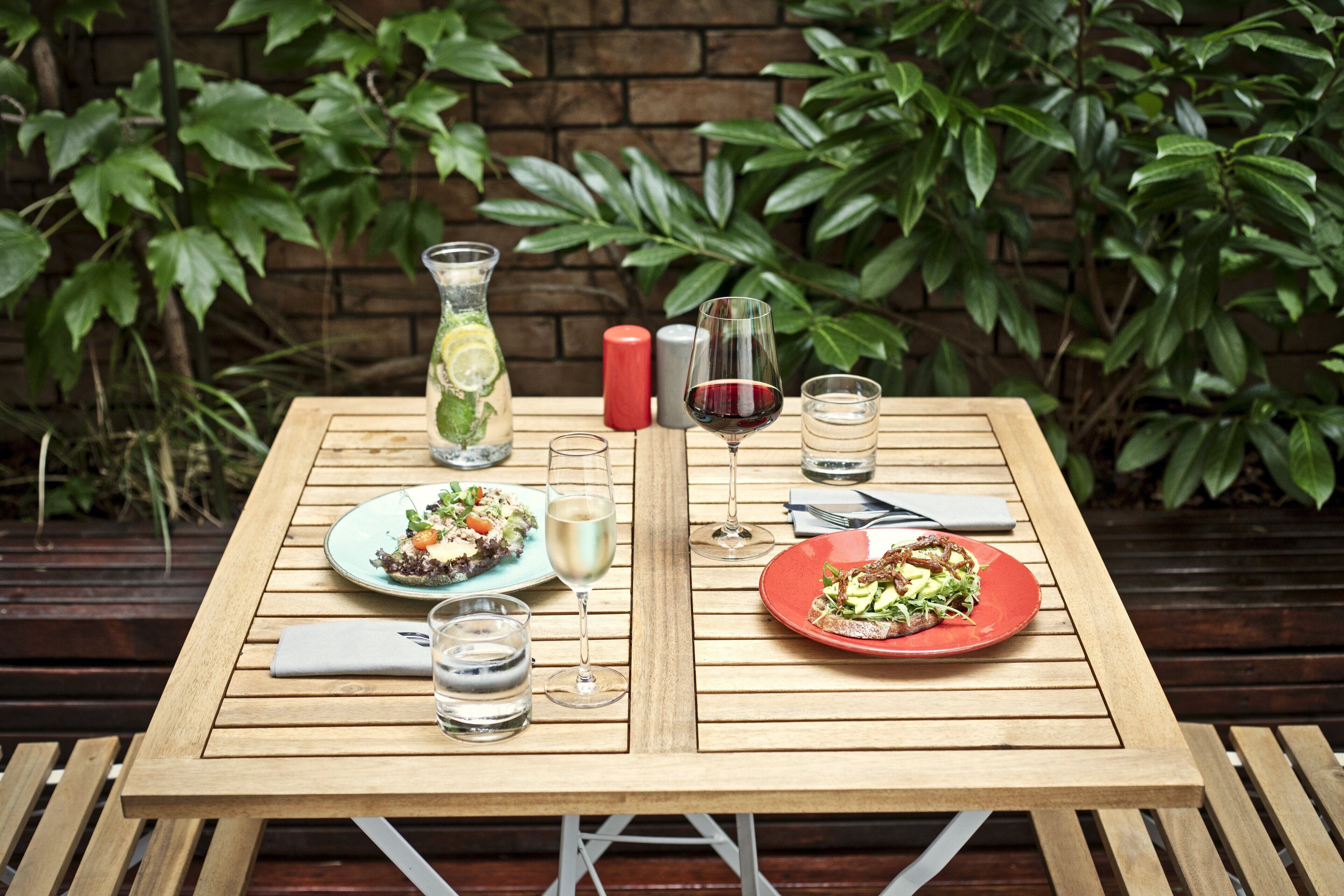 A wooden table set with two plates of food, two glasses of wine, a vase with a drink, and salt and pepper shakers.