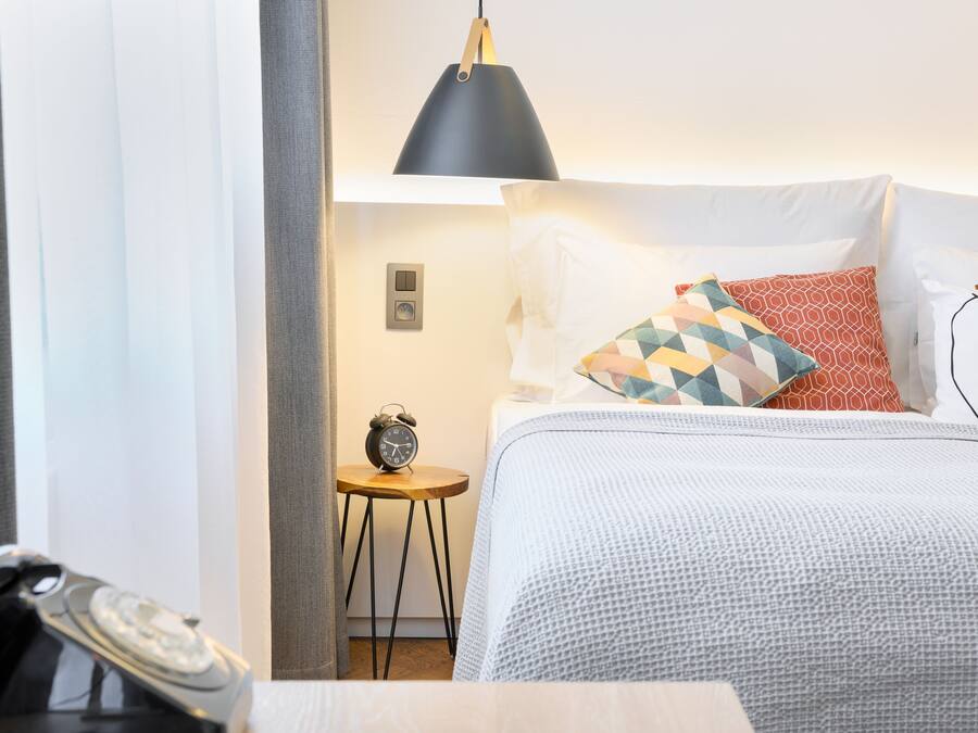 A neatly made bed with patterned pillows, a bedside table with a lamp, and a classic alarm clock.