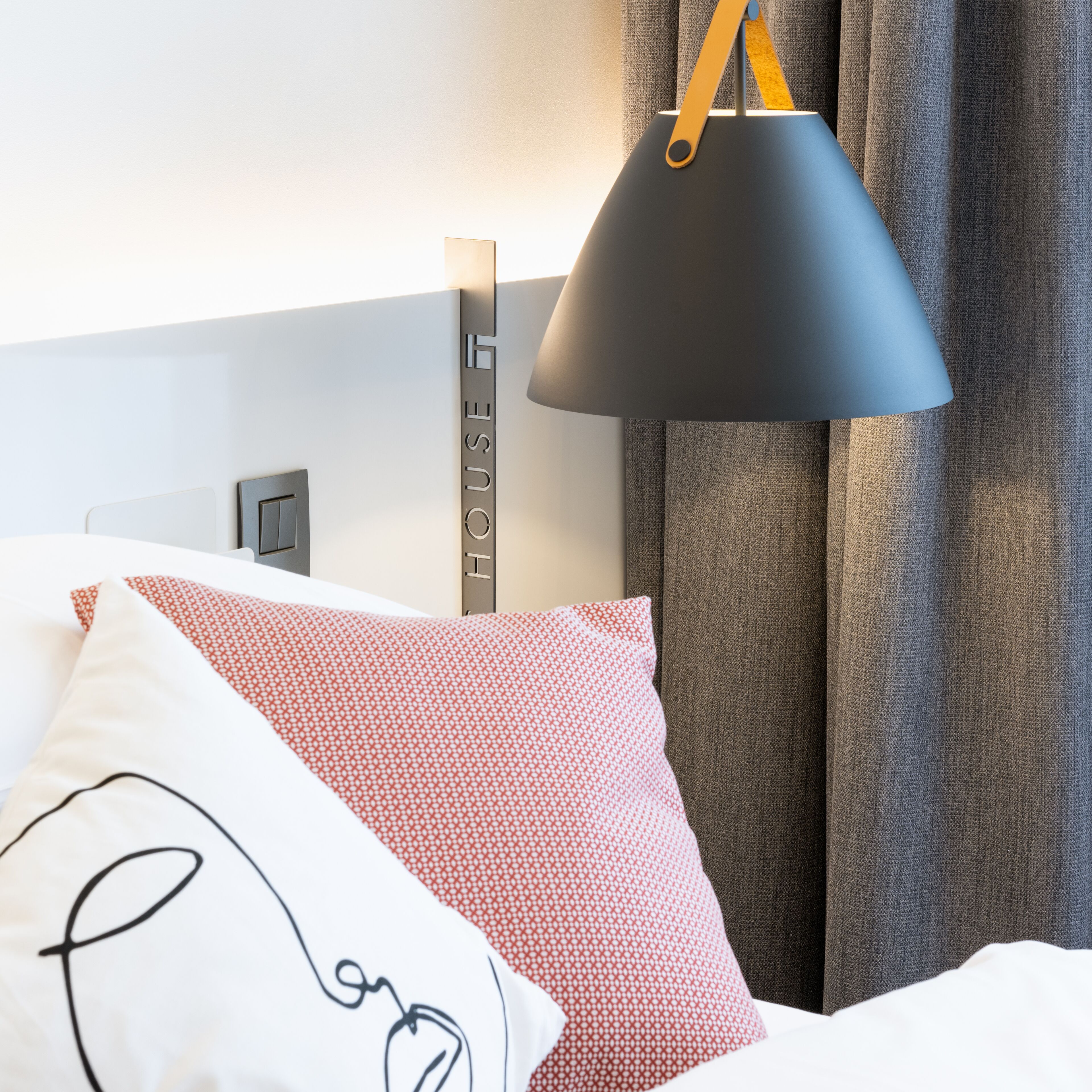 Una cama bien tendida con almohadas estampadas, una lámpara de noche y un cartel en la pared que dice 'HOUSE'.