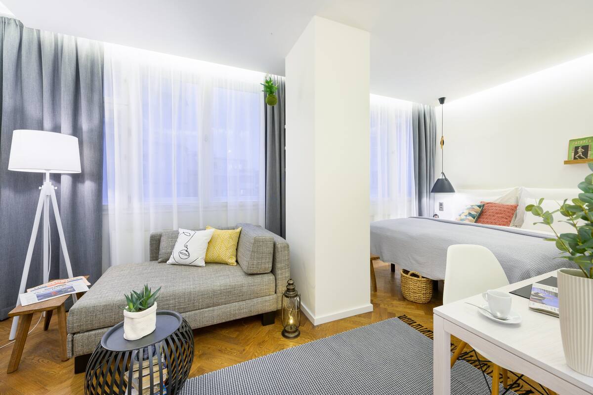 A modern hotel room with a sofa, a small table with a lamp, a bed, and a desk with a cup and a plant.