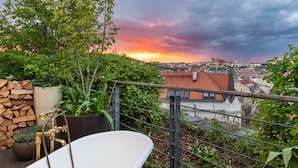 A rooftop terrace with a white bathtub, a wooden deck, and a view of a cityscape during sunset.