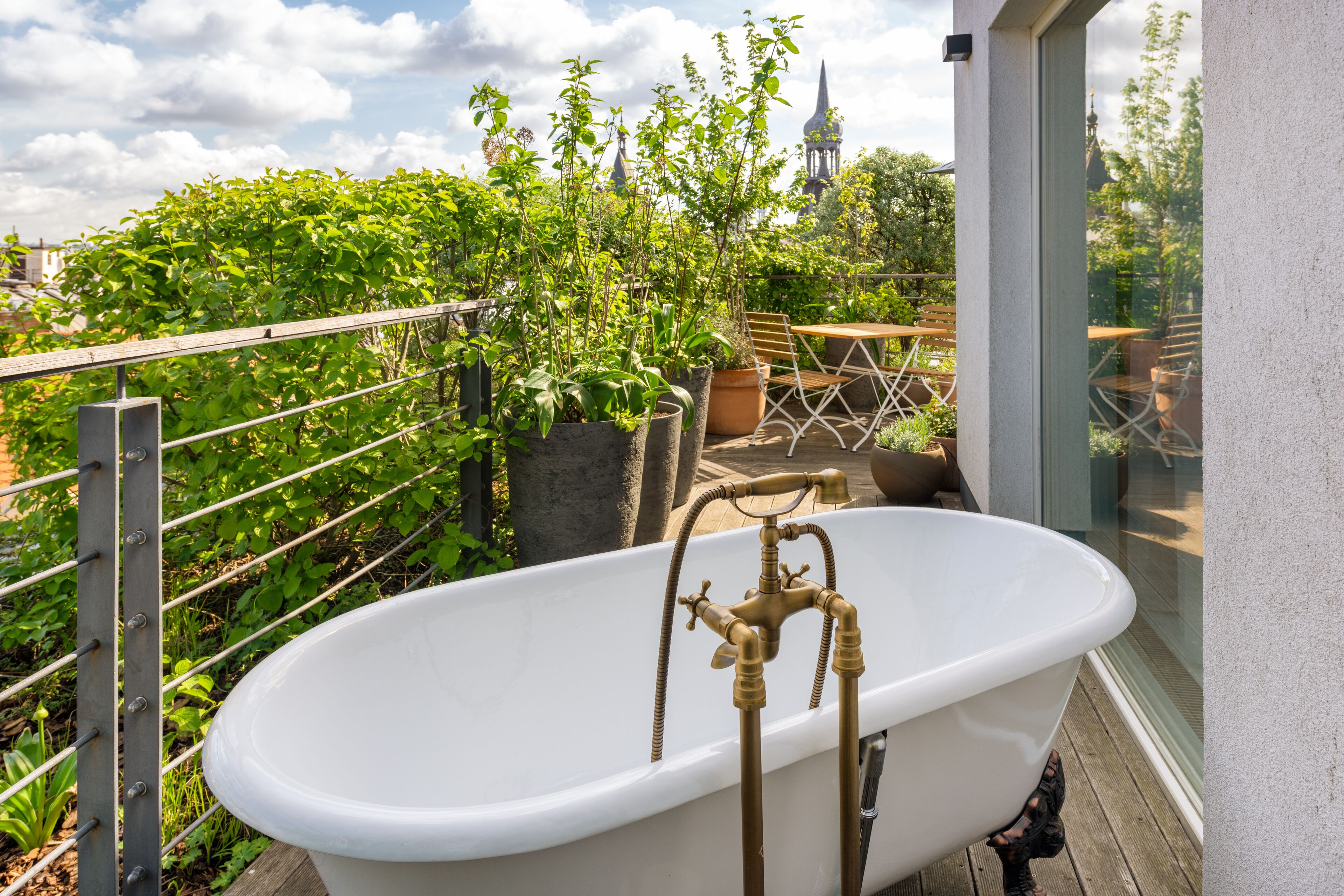 A white freestanding bathtub with brass fixtures on a balcony with a view of a church spire.