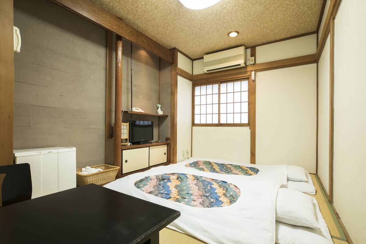 Una habitación tradicional japonesa con suelo de tatami, futón, televisor pequeño y refrigerador.