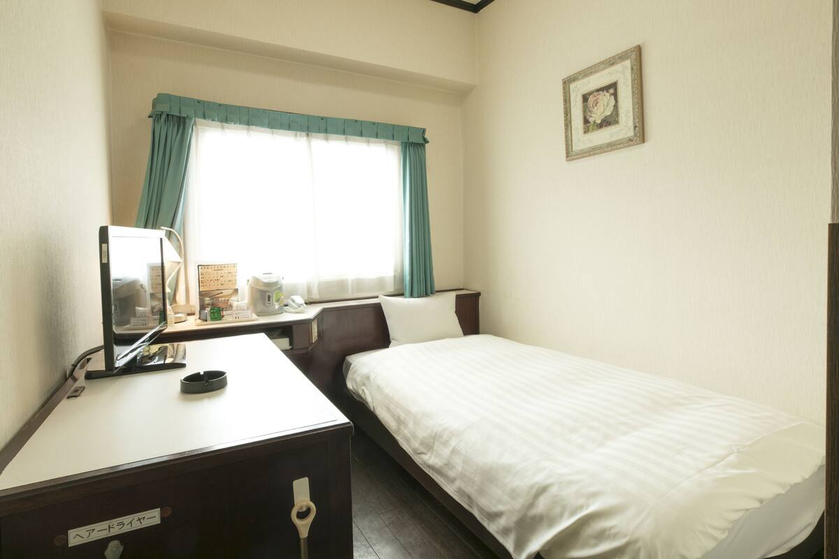 Una habitación de hotel con una cama individual, un escritorio con una computadora, una mesita con un teléfono y una ventana con cortinas.