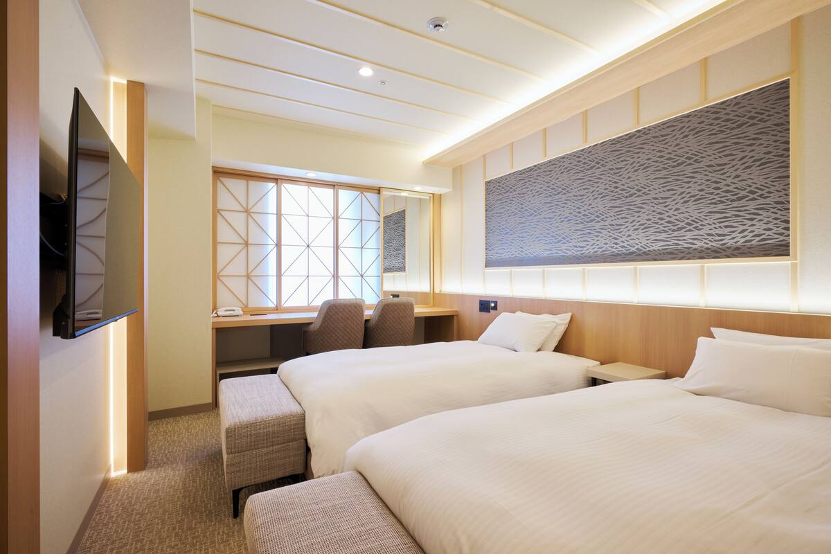 A hotel room with two beds, a desk with a chair, a TV mounted on the wall, and a large window with a view.