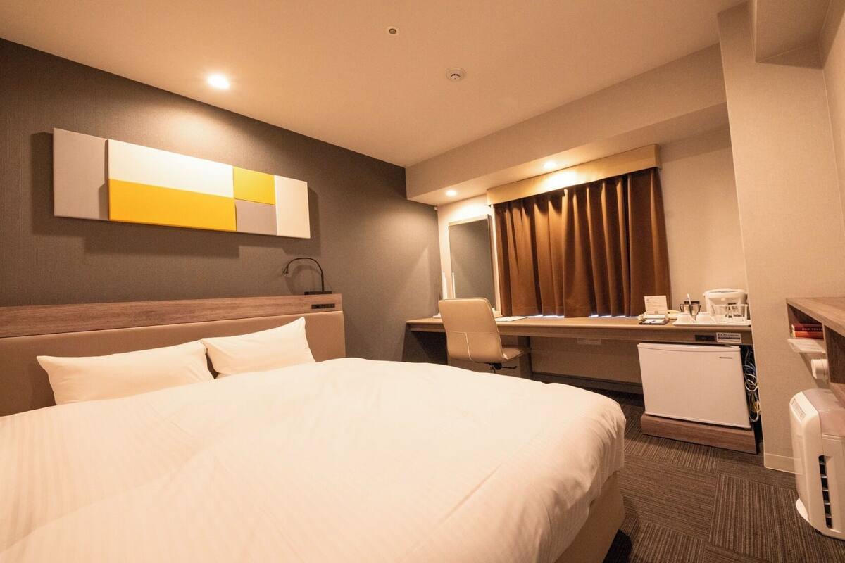 A hotel room with a bed, a desk with a chair, a small refrigerator, a coffee maker, and a television.
