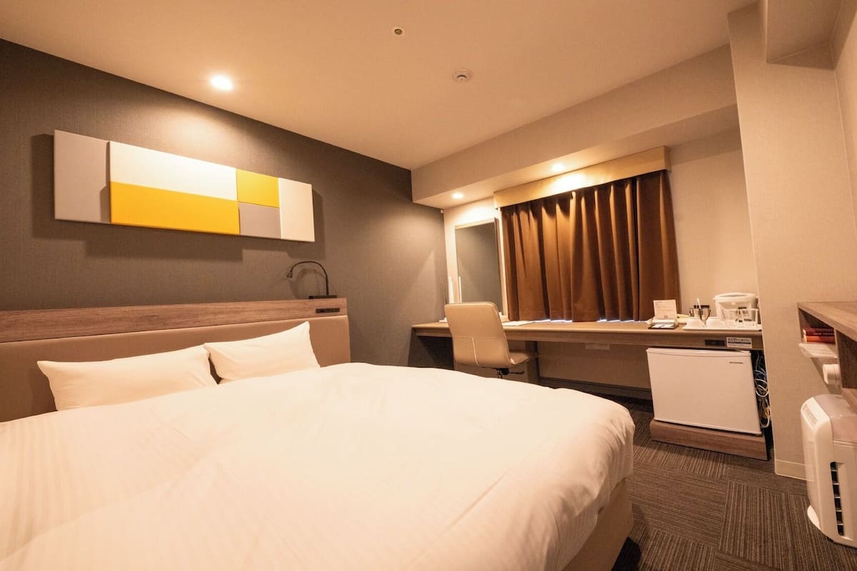 A hotel room with a bed, a desk with a chair, a small refrigerator, a coffee maker, and a television.