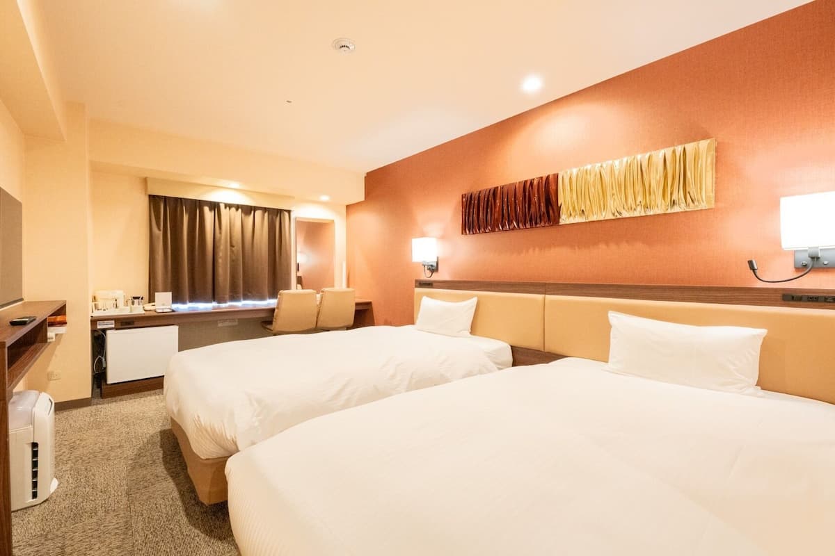 A hotel room with two beds, a desk with a flat-screen TV, a desk lamp, and a wall-mounted light.