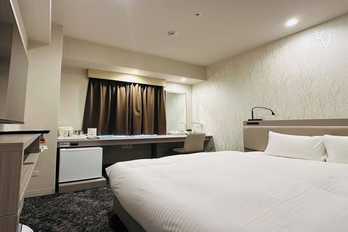 A hotel room with a bed, a desk, a television, a mini-fridge, and a bathroom mirror.