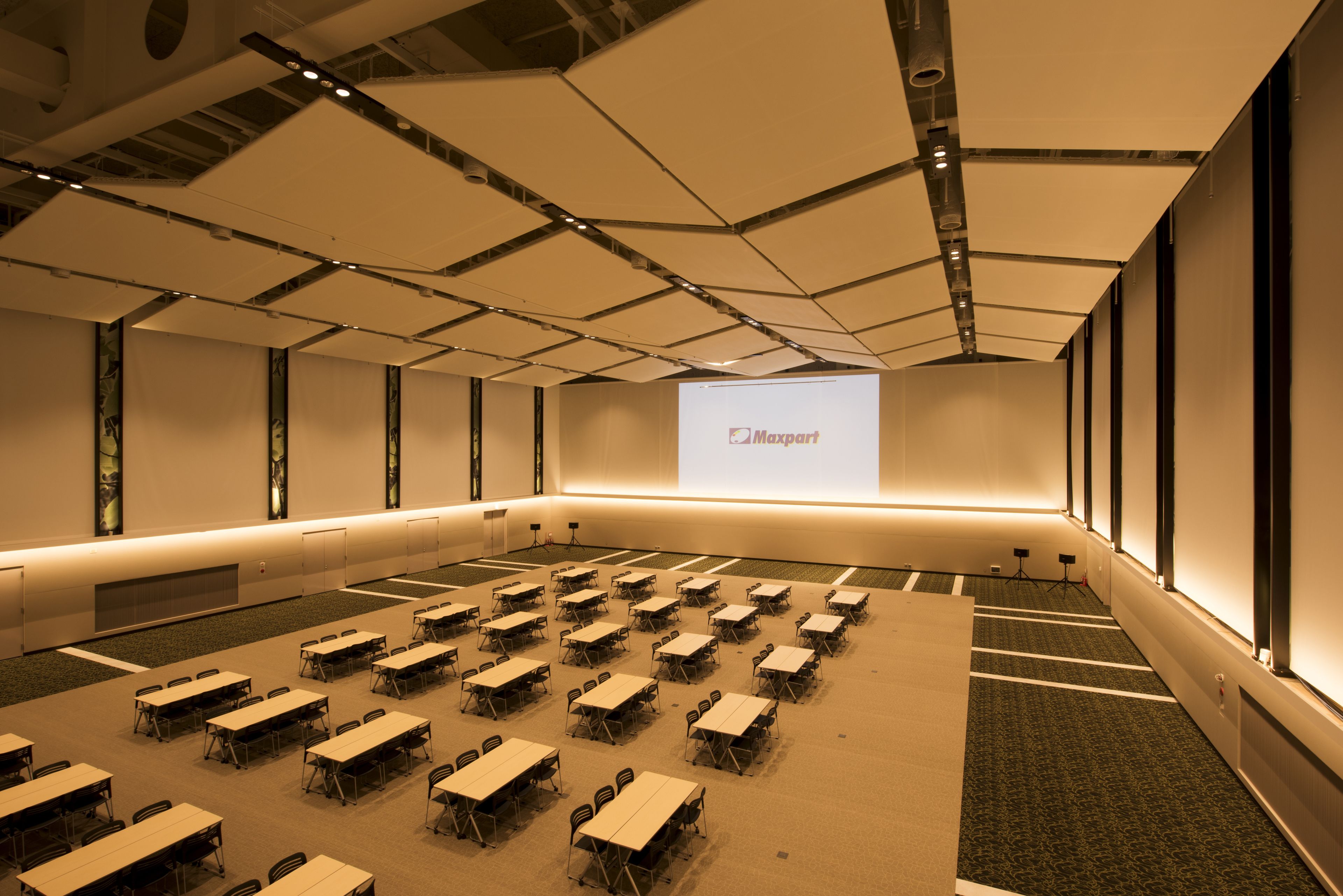 A conference room with multiple tables and chairs, a large screen displaying 'Maxpart', and a high ceiling with exposed beams.