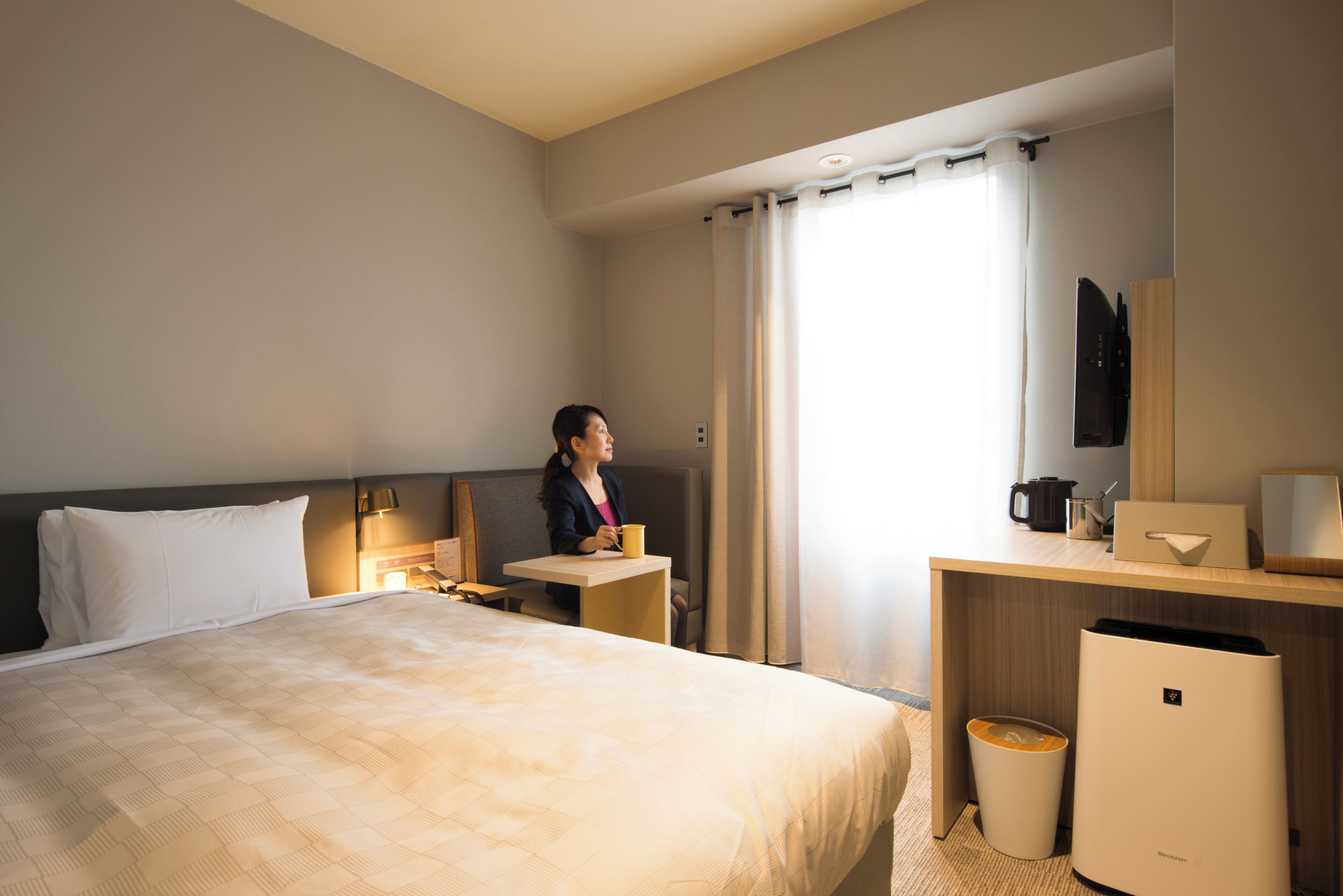 Habitación de hotel con cama, escritorio, silla, cafetera, cesto de basura y espejo.