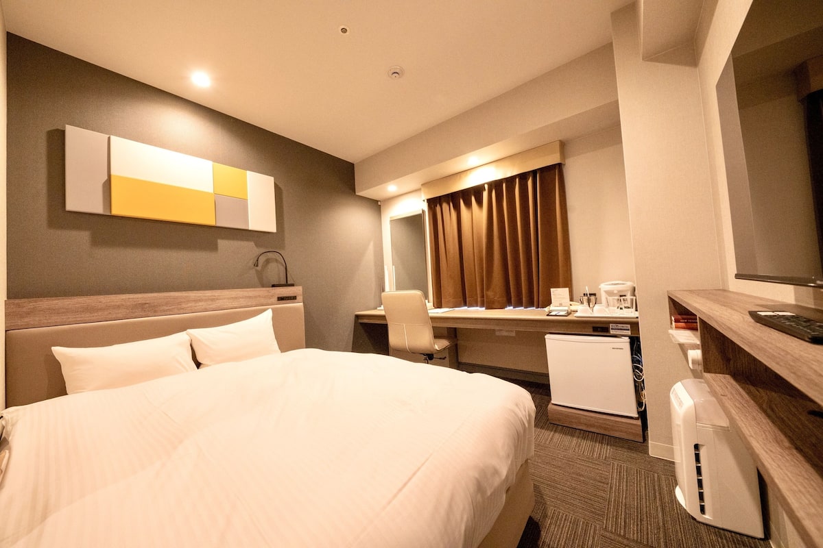 A hotel room with a bed, desk, chair, and a small kitchenette.