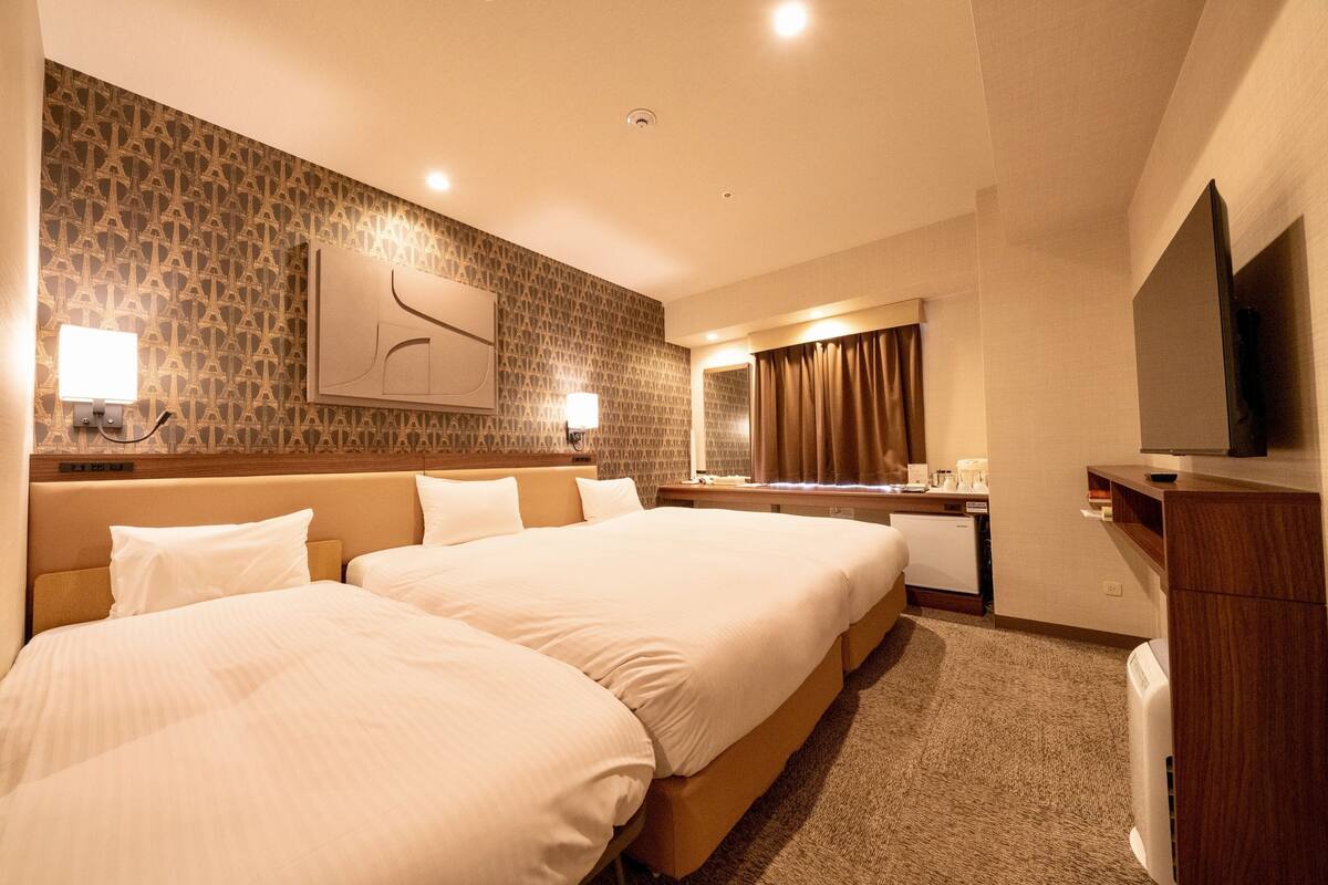 A hotel room with two beds, a flat-screen TV, a desk with a chair, a mirror, and a wall-mounted lamp.