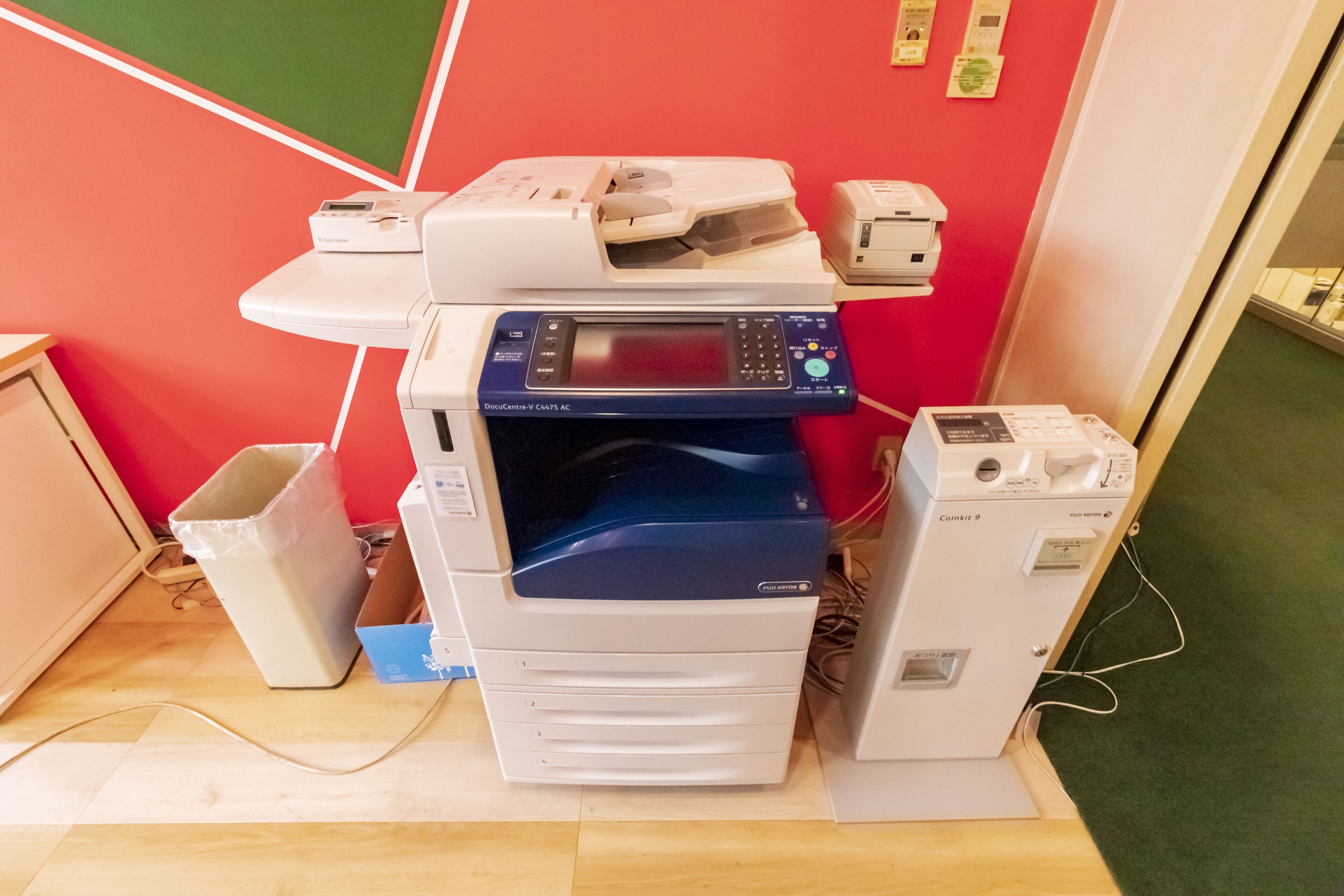 Ein Multifunktionsdrucker auf einem Schreibtisch zusammen mit einem Papierkorb und einem kleinen Tisch mit einem Telefon.