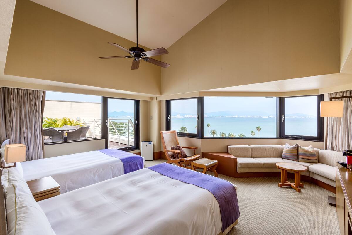 A hotel room with two beds, a sofa, a chair, a small table, and a ceiling fan.