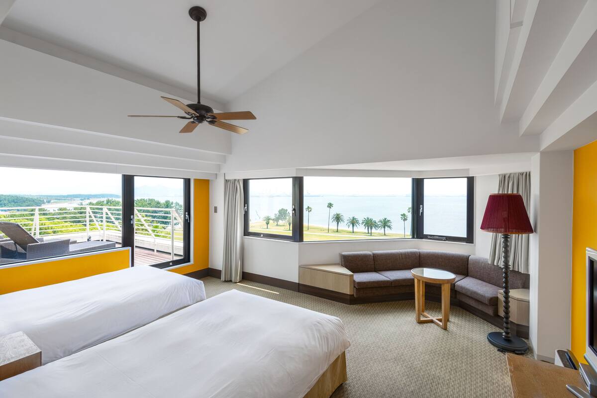 A hotel room with two beds, a sofa, a small table, and a lamp. There are large windows offering a view of the ocean and palm trees.