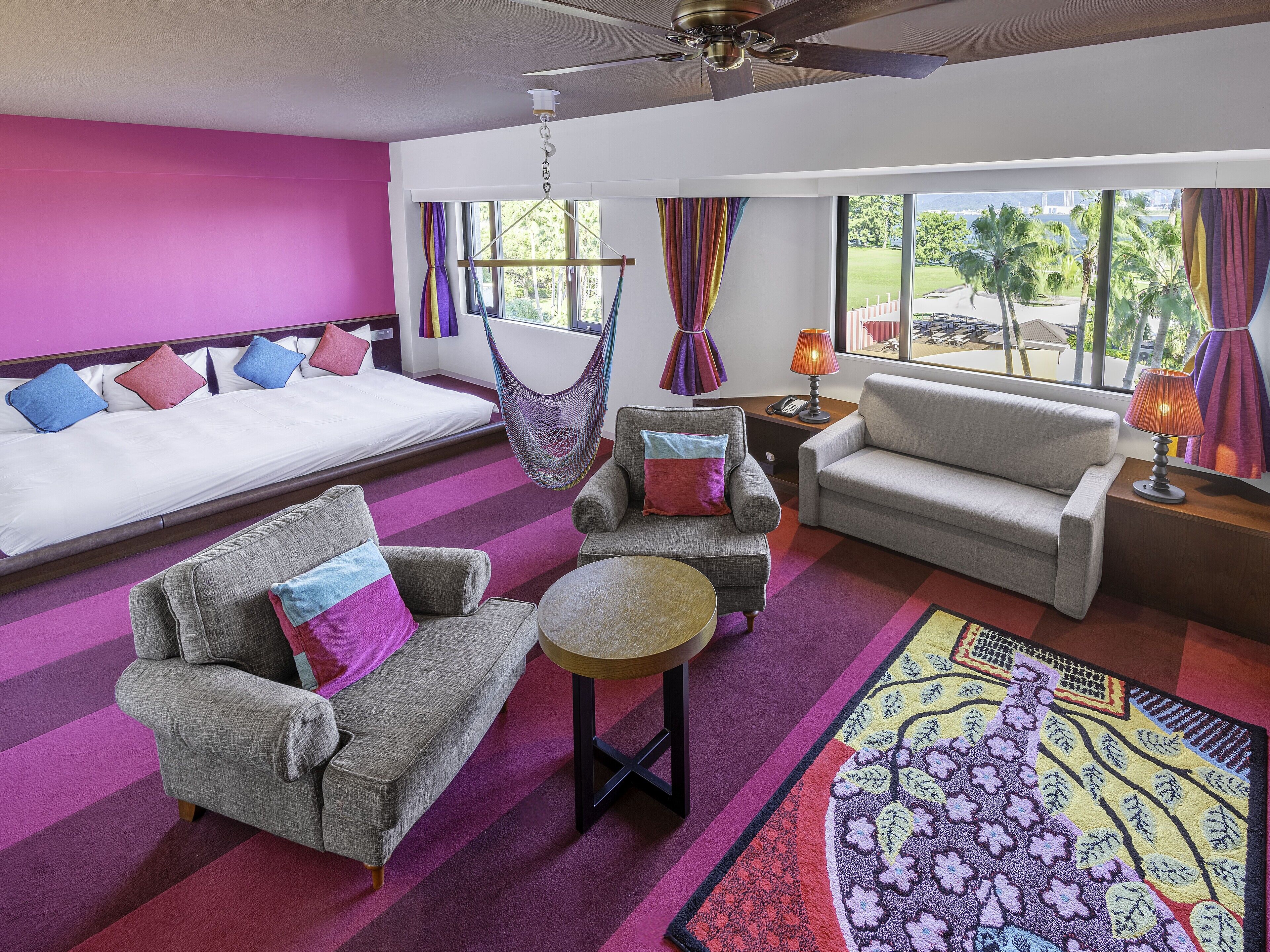 A hotel room with a bed, two sofas, a chair, a small table, a hanging chair, and a large window with a view of palm trees and a bridge.