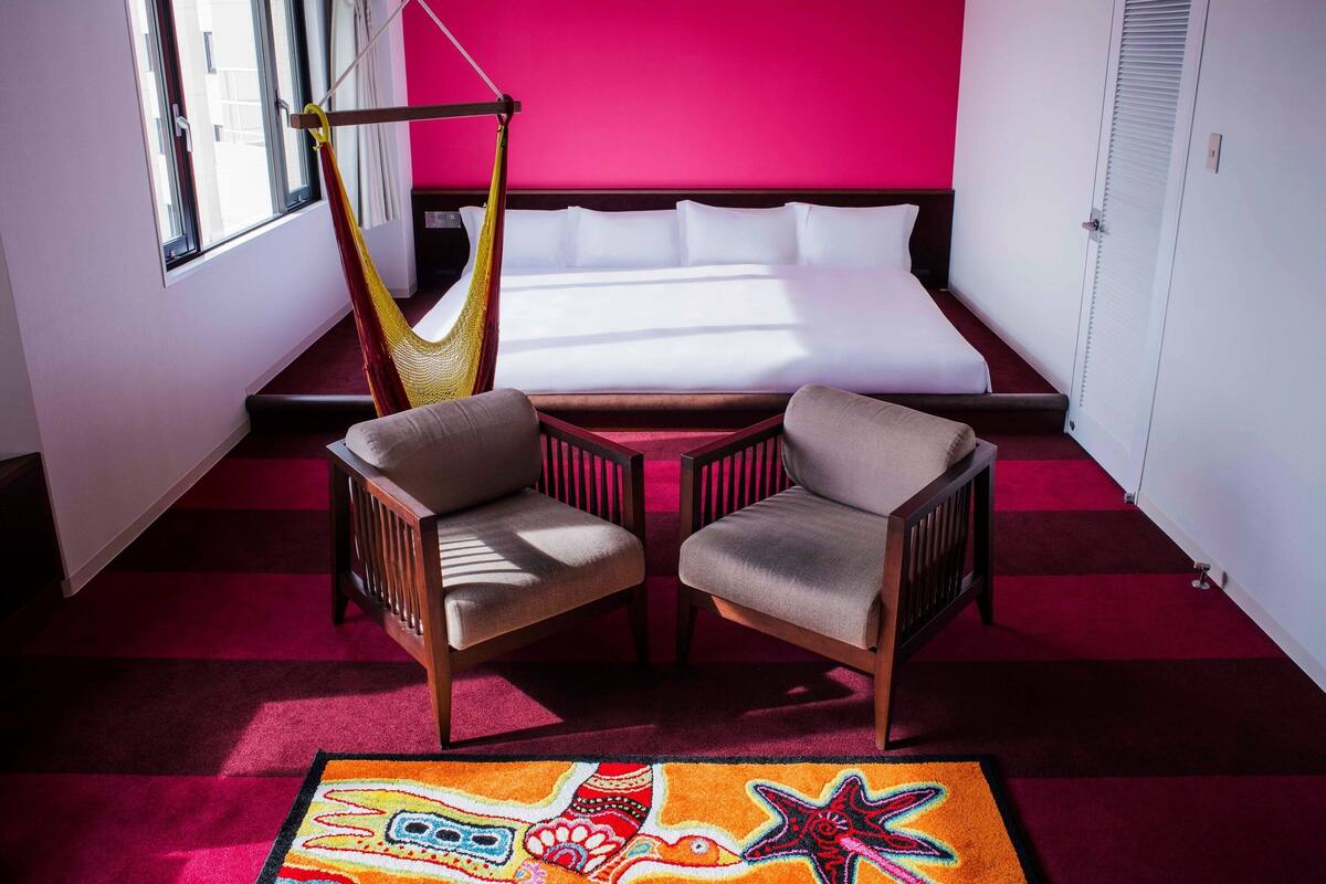 A hotel room with a bed, two chairs, a hammock, a colorful rug, and a window.