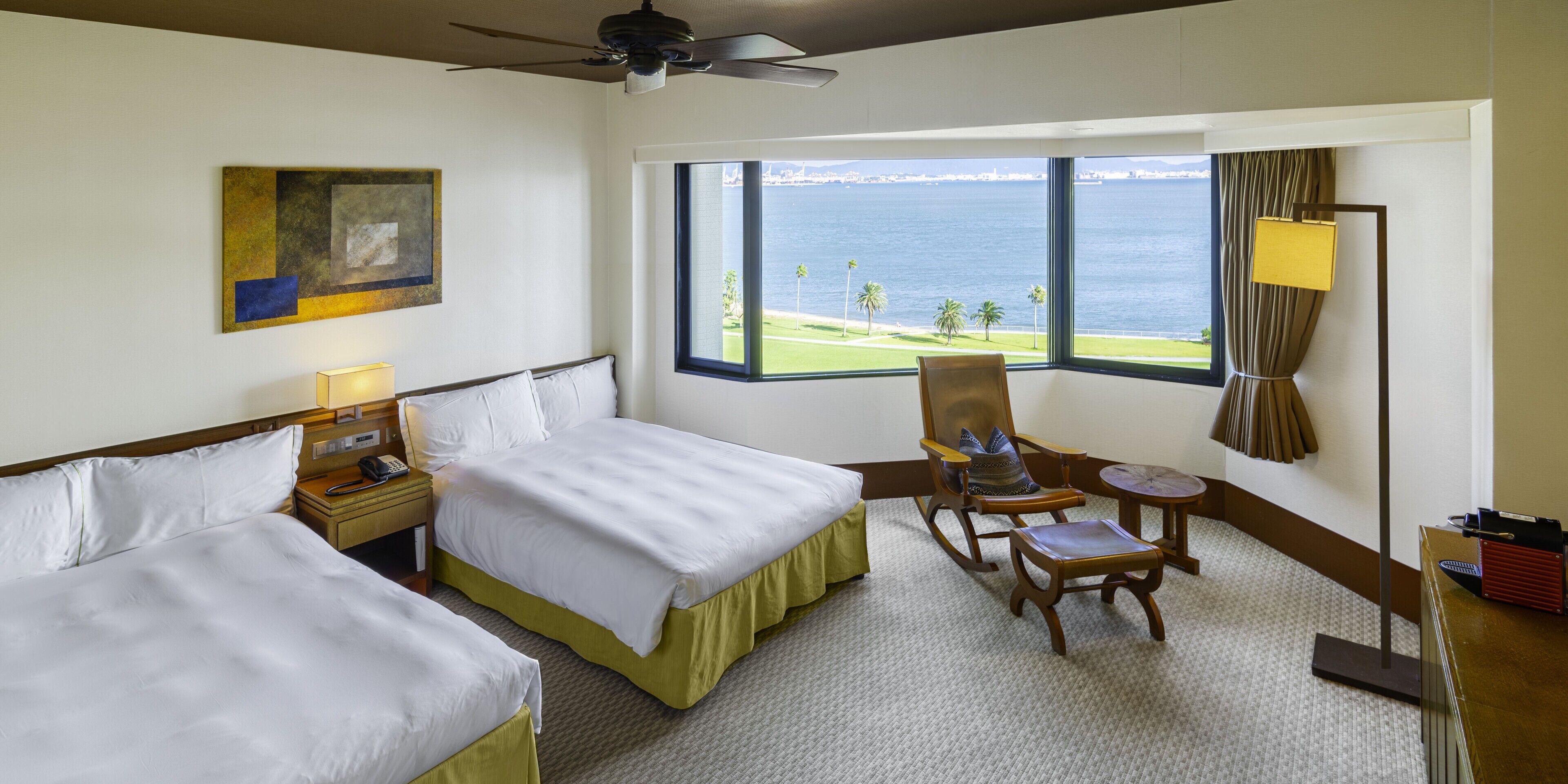 A hotel room with two beds, a desk, a chair, a large window with a view of the sea, and a ceiling fan.