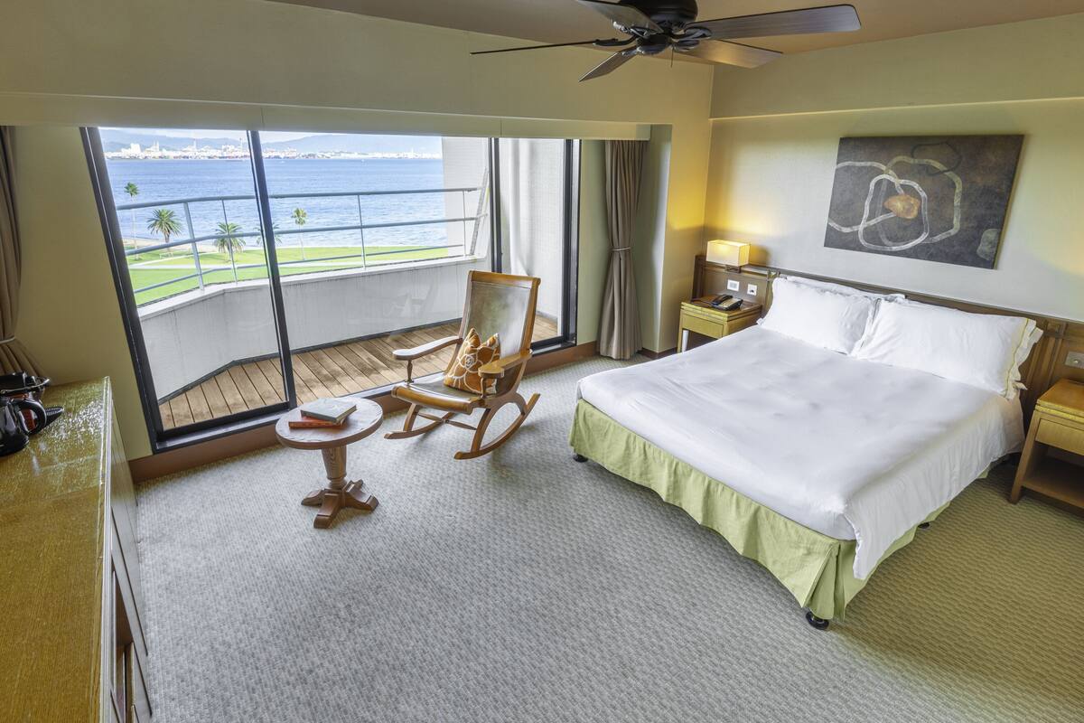 A hotel room with a large bed, a rocking chair, a small table, and a view of the sea through large windows.