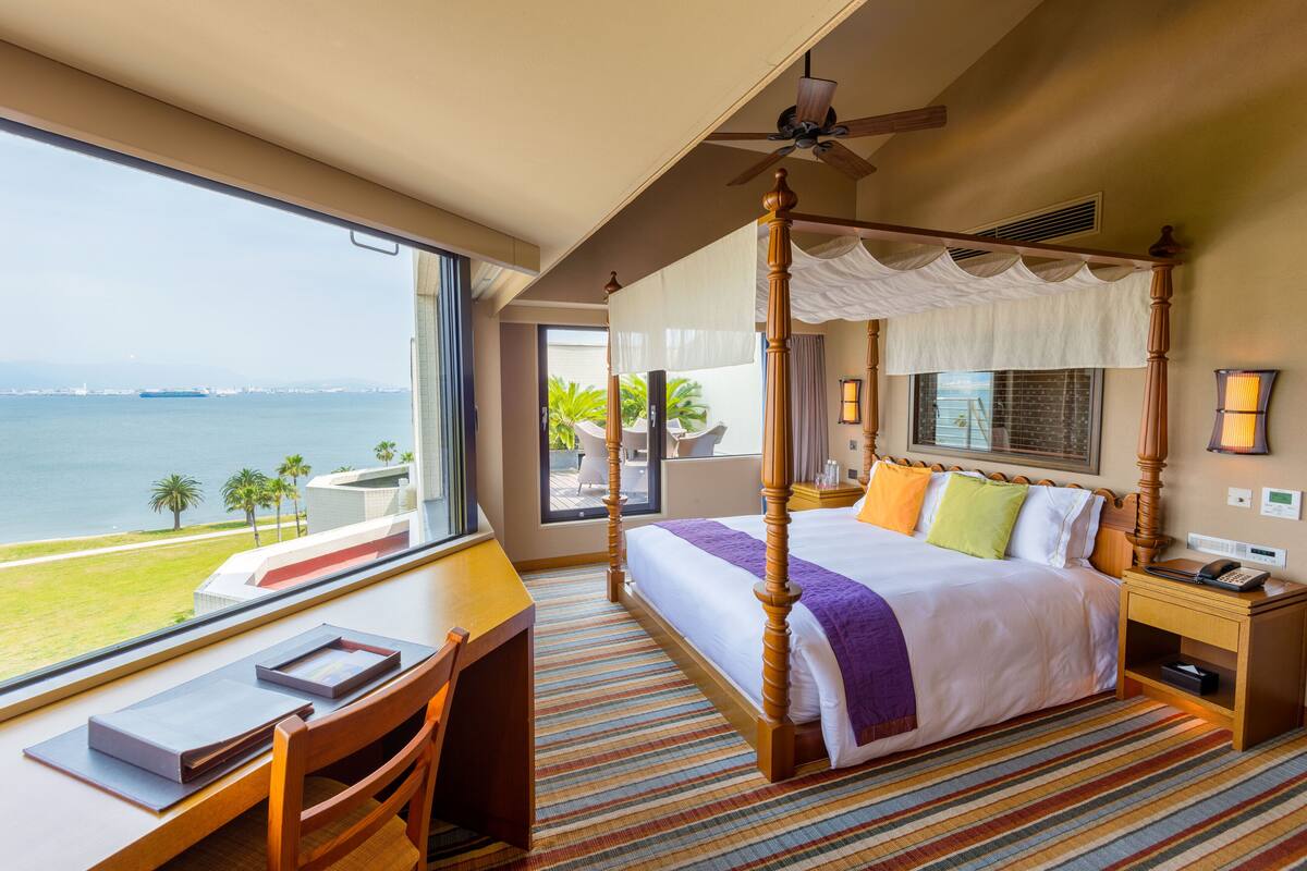 A four-poster bed with a canopy, a desk with a chair, and a view of the sea.