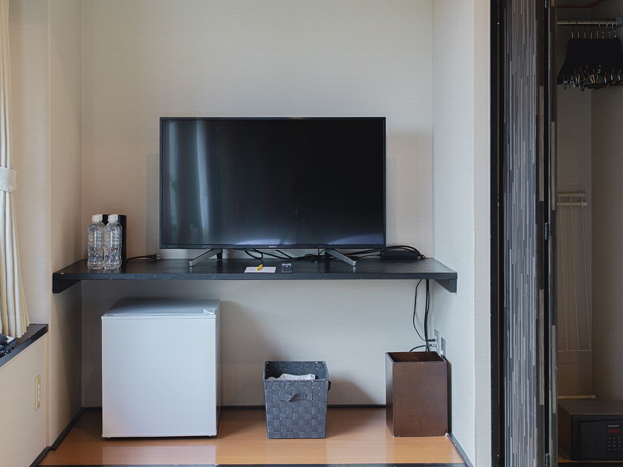 Ein kompakter Wohnbereich mit Flachbildfernseher an einer dunkelgrauen Wand, einem kleinen weißen Kühlschrank, einer grauen Aufbewahrungsbox und einem Holzschrank.