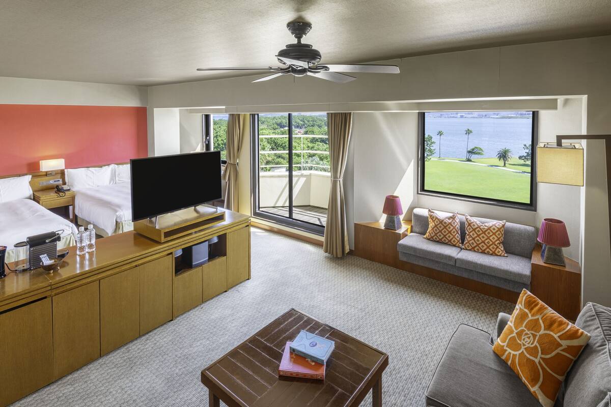 A hotel room with a TV, a sofa, a coffee table, and a balcony with a view of the water.