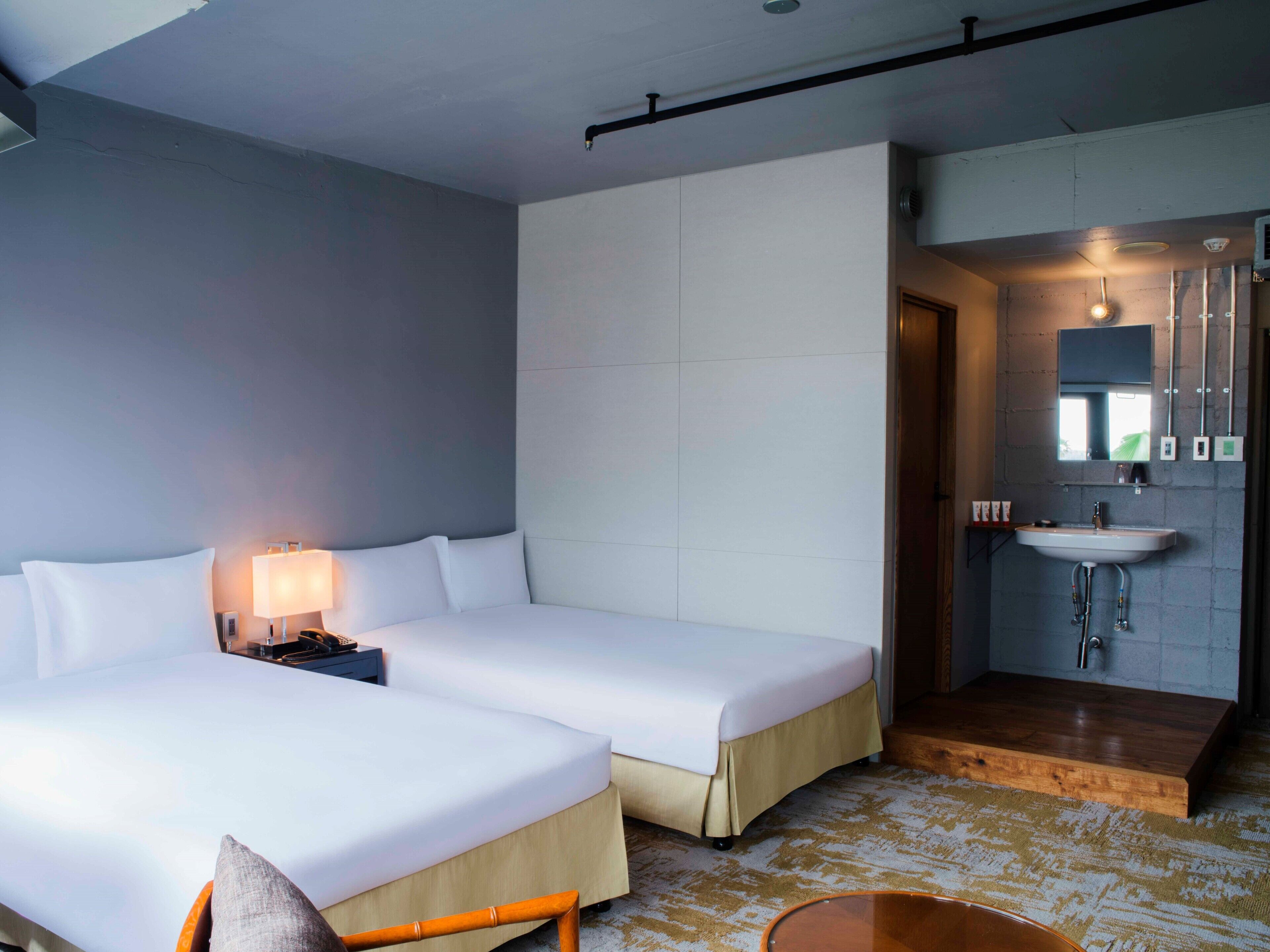 A hotel room with two beds, a bedside table with a lamp, a wooden floor, and a bathroom with a sink and mirror.